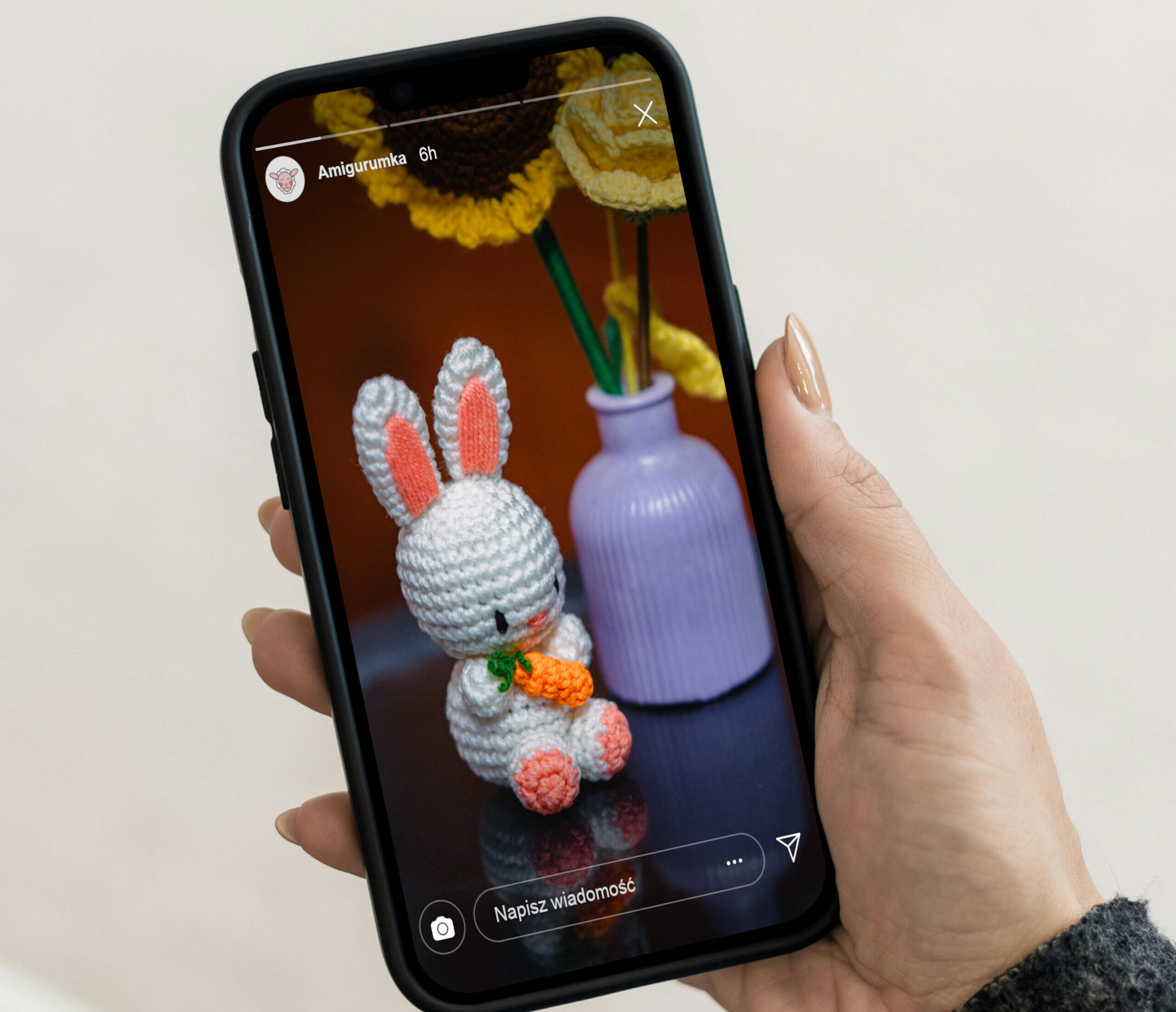 Kobieta trzyma telefon w jednej dłoni. Na ekranie smartfona jest wizualizacja zdjęcia profilowego na realcji Instagramowej Amigurumki.